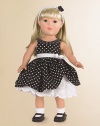 This 18 inch play doll has blue eyes and ivory colored, side-parted, shoulder length hair. She wears a sleeveless dress of black with white polka dots. The left side of the skirt, of the dress, is gathered up to reveal a white cotton underskirt that has flower appliqués and a scalloped hem.